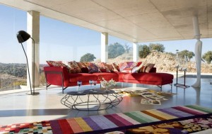 Cozy-Living-Room-With-L-Shaped-Red-Sofa-Many-Roche-Bobois-Cushions-Unusual-Table-And-Colorful-Carpet